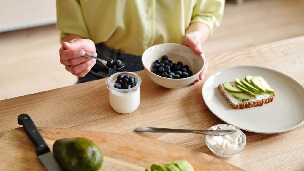A person prepares a healthy breakfast with yogurt, blueberries, and avocado toast.