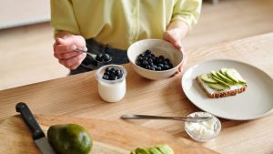 A person prepares a healthy breakfast with yogurt, blueberries, and avocado toast.